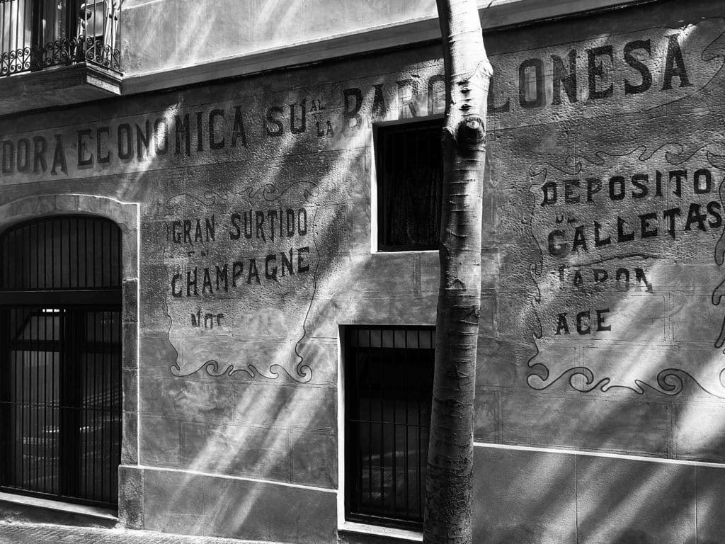 Walking along Carrer Margarit today, echoes of the past reappear in subtle ways. Recent plaster removal revealed faded signage of a former corner inn, one of the many places once famed for champagne, imported wines, and liqueurs; like the walls themselves deciding to speak again.